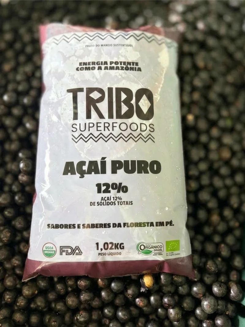 Embalagem de 1,02 kg de Açaí Puro 12% de sólidos totais da Tribo Superfoods, com selos orgânicos, apoiada sobre frutos de açaí recém-colhidos